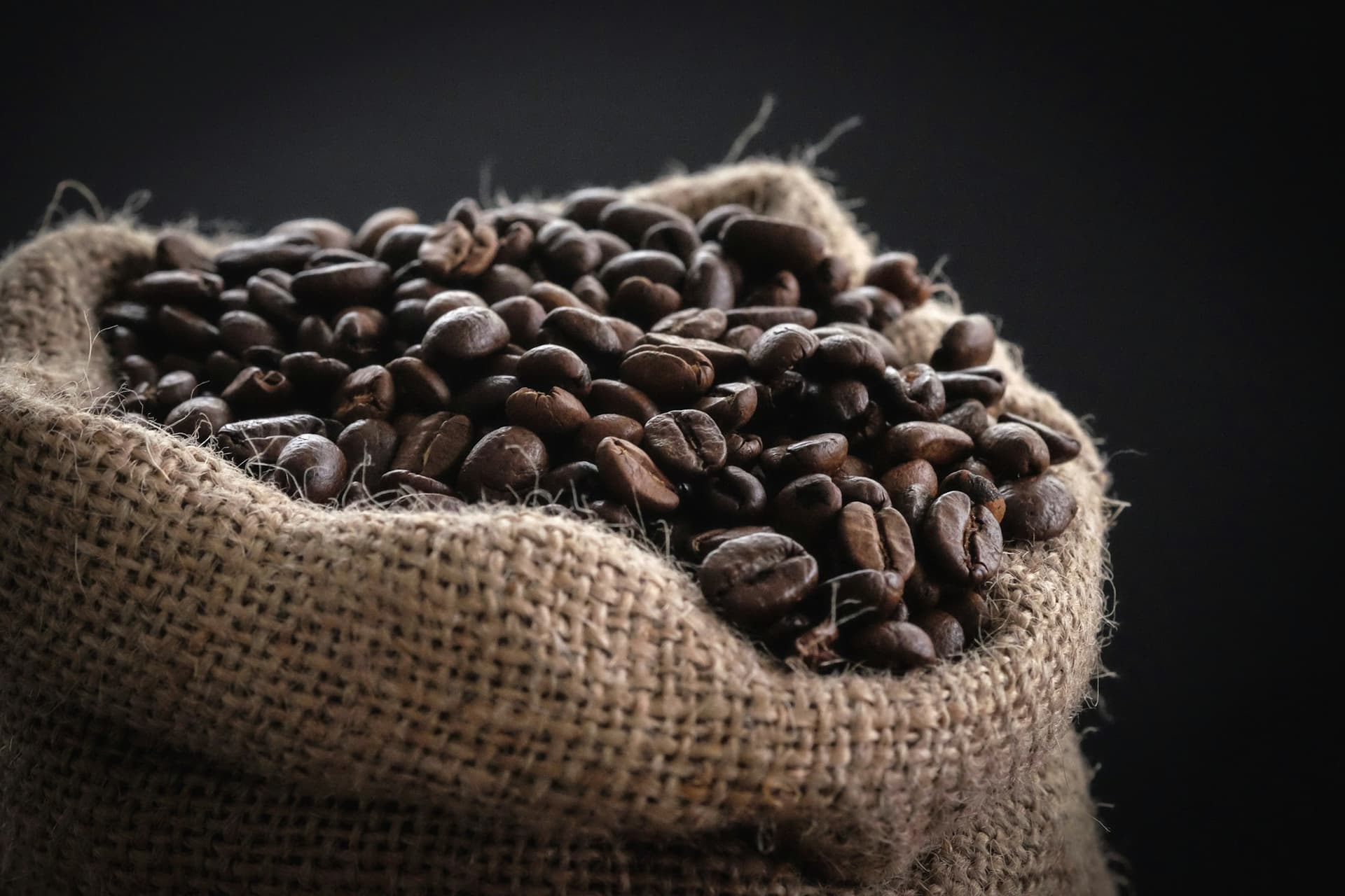An open jute sack filled to the mouth with roasted coffee beans, against a dark background.