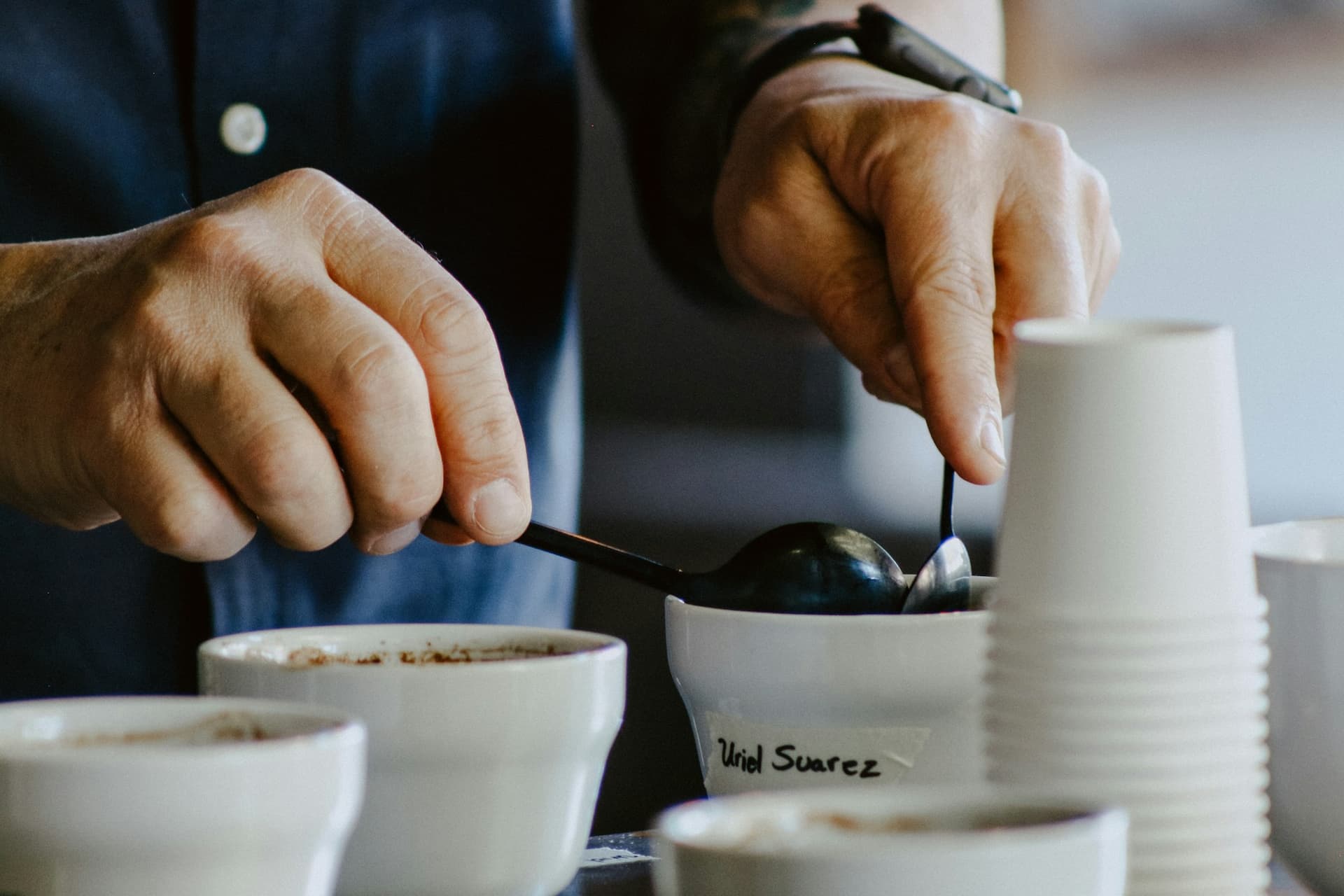 A cupping table in use: two hands breaking the crust with cupping spoons across coded sample bowls.