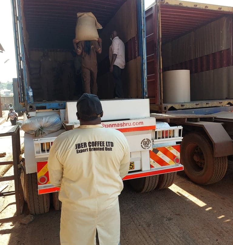 Workers carrying jute coffee sacks into a shipping container, one in overalls marked Jber Coffee Ltd.