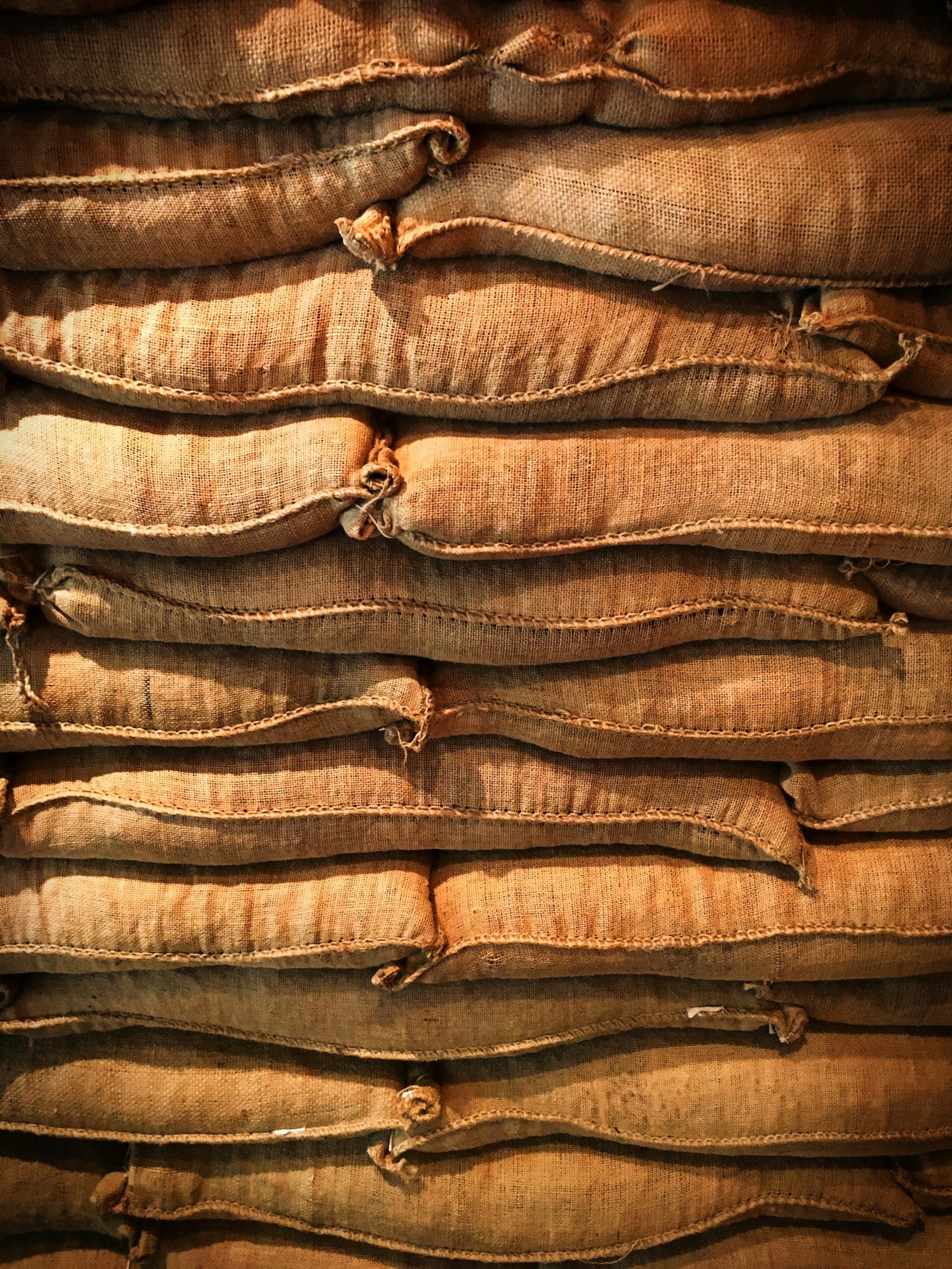 Filled jute sacks stacked in courses, each hand-stitched closed along the mouth, the weave and the seam running the width of the frame.