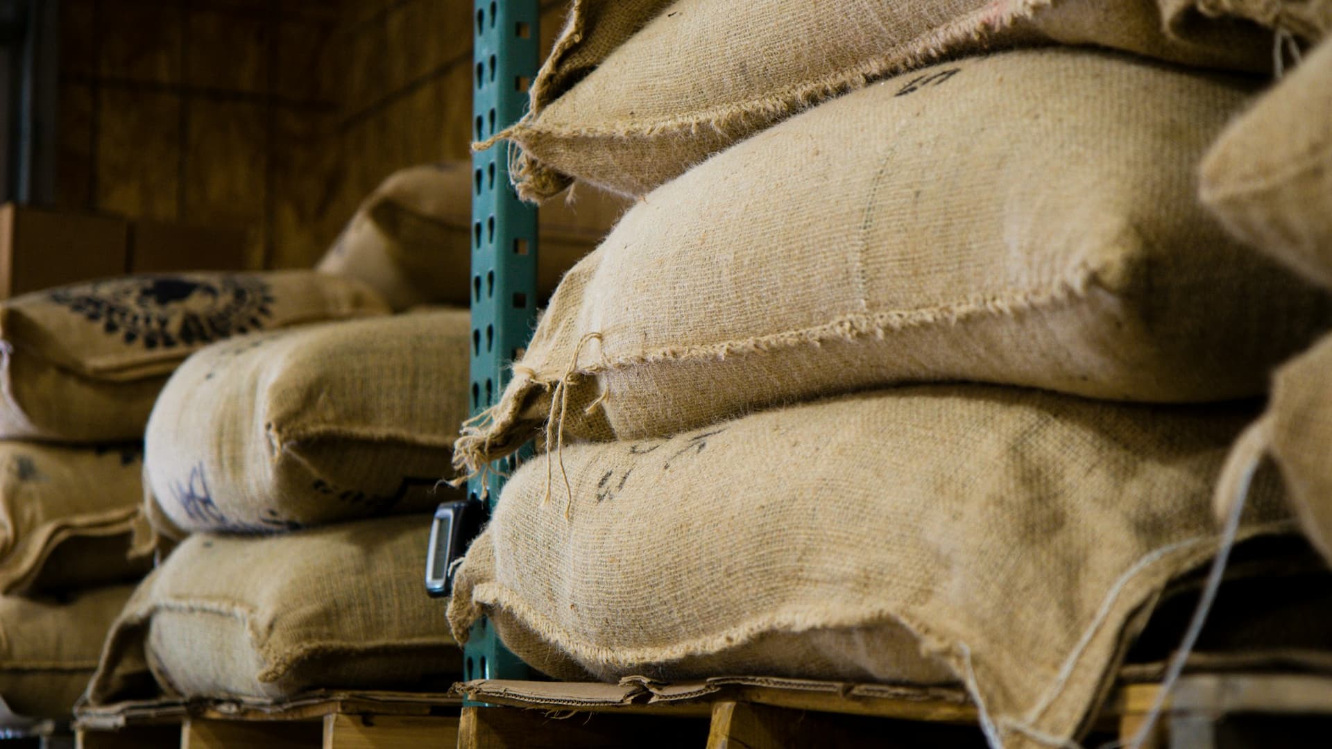 Jute coffee sacks stacked on wooden pallets in racking inside a warehouse, tied at the neck with twine.