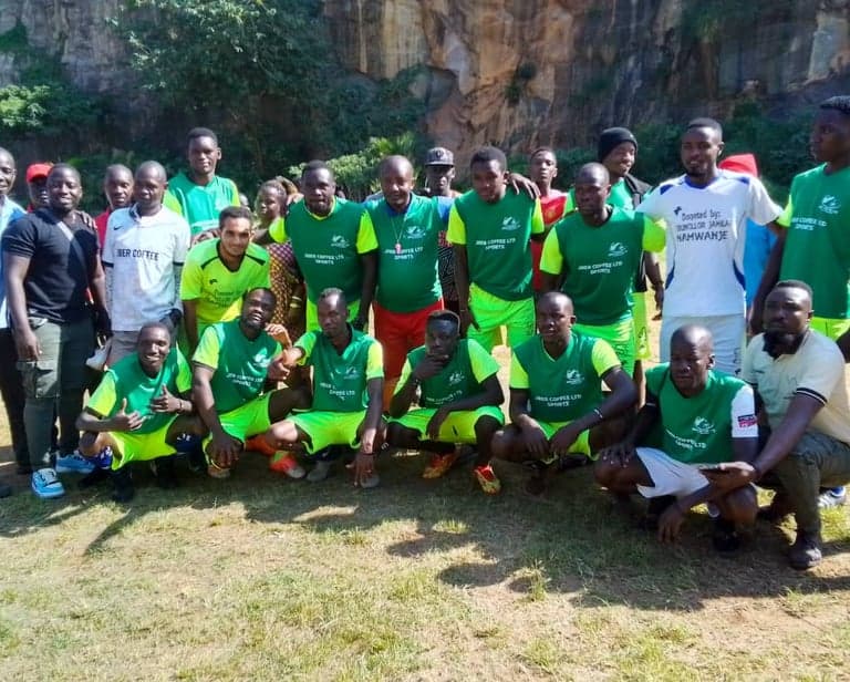 The Jber Coffee Ltd Sports squad photographed at a pitch, players in green and yellow kit with supporters behind.